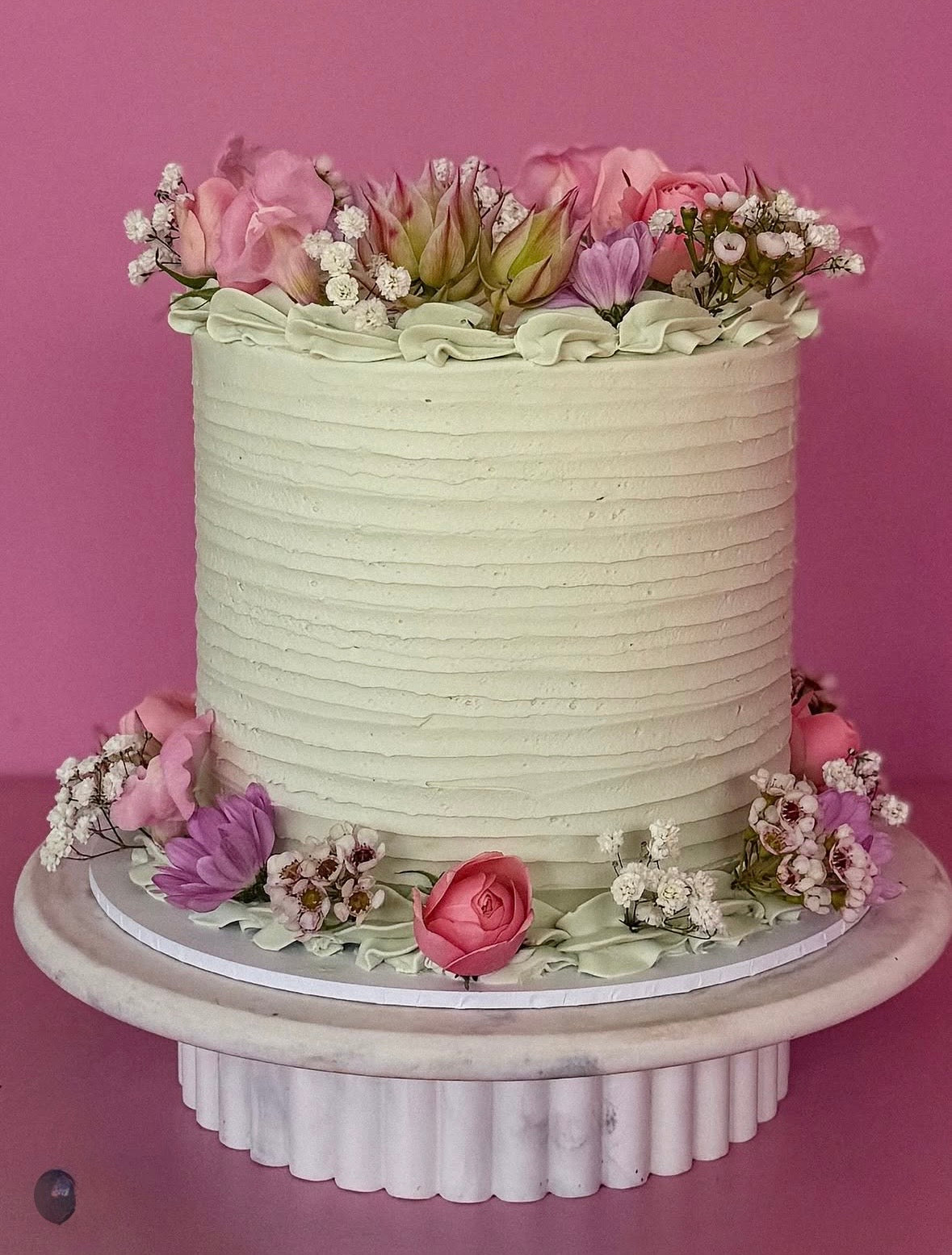 A single-tier round cake with textured off-white frosting, decorated with pink and purple flowers and green leaves, sitting on a white fluted cake stand against a solid pink background.