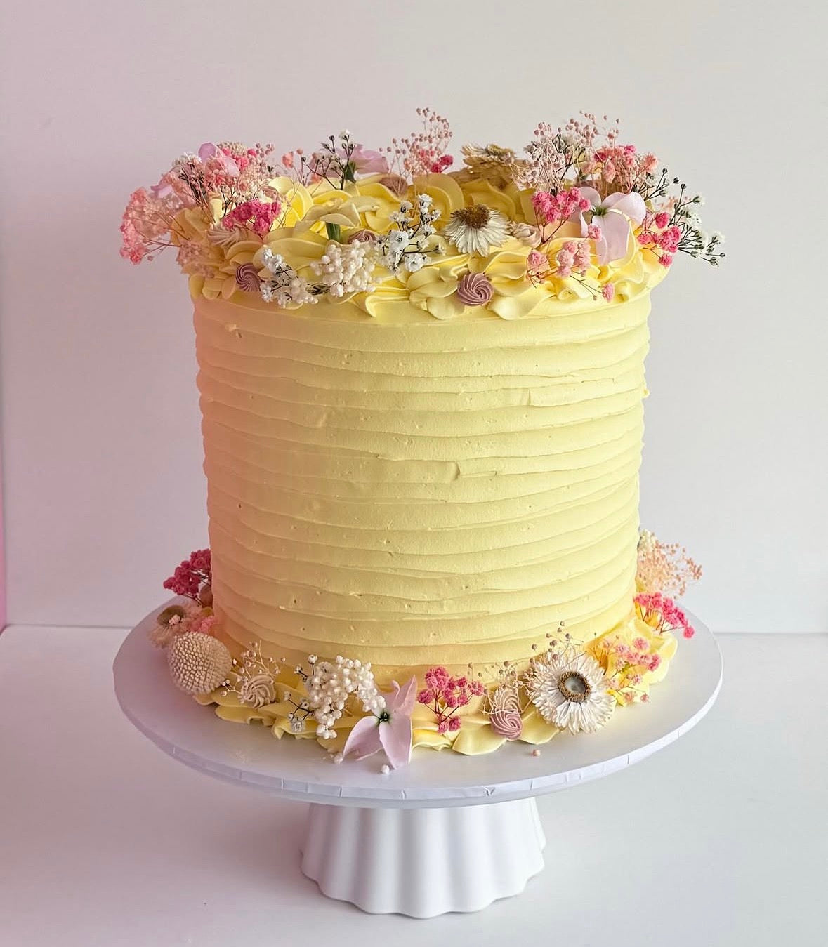 A pale yellow frosted cake with textured horizontal ridges, decorated with pink and yellow flowers and dried botanicals, displayed on a white pedestal stand against a light pink and white background.
