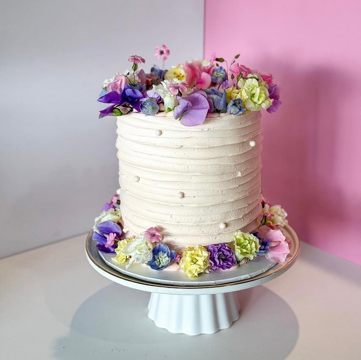 A single-tier round cake with white ruffled frosting, decorated with colorful artificial flowers in purple, pink, yellow, and green, displayed on a white pedestal stand against a split white and pink background.
