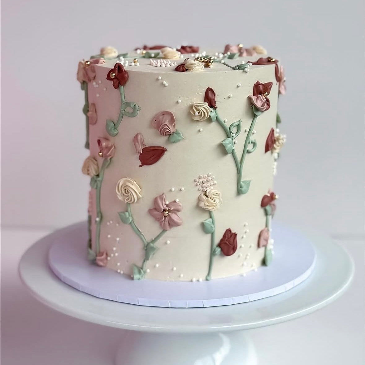 A round frosted cake with delicate piped floral decorations in pink, cream, and burgundy, displayed on a white cake stand against a soft light gray background.