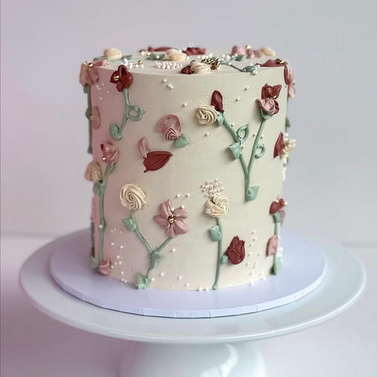 A round frosted cake with delicate piped floral decorations in pink, cream, and burgundy, displayed on a white cake stand against a soft light gray background.