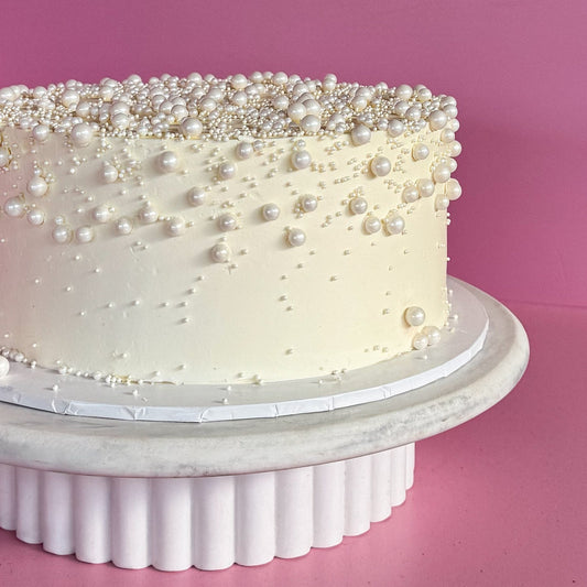 A round cake with smooth ivory frosting, decorated with scattered white pearl-like beads and sprinkles, displayed on a white fluted pedestal stand against a solid pink background.