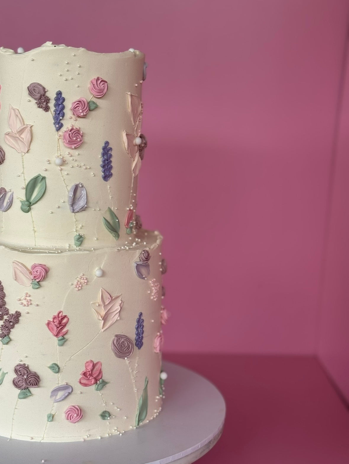 A two-tiered cream cake decorated with colorful piped flowers, leaves, and small white pearls, displayed on a white cake stand against a solid pink background.